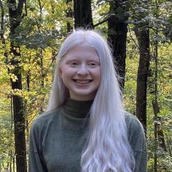Sarah smiles at the camera in front of a background of green trees. She has albinism, and as a result her long hair is white-blonde. She is wearing a green high-neck sweater.